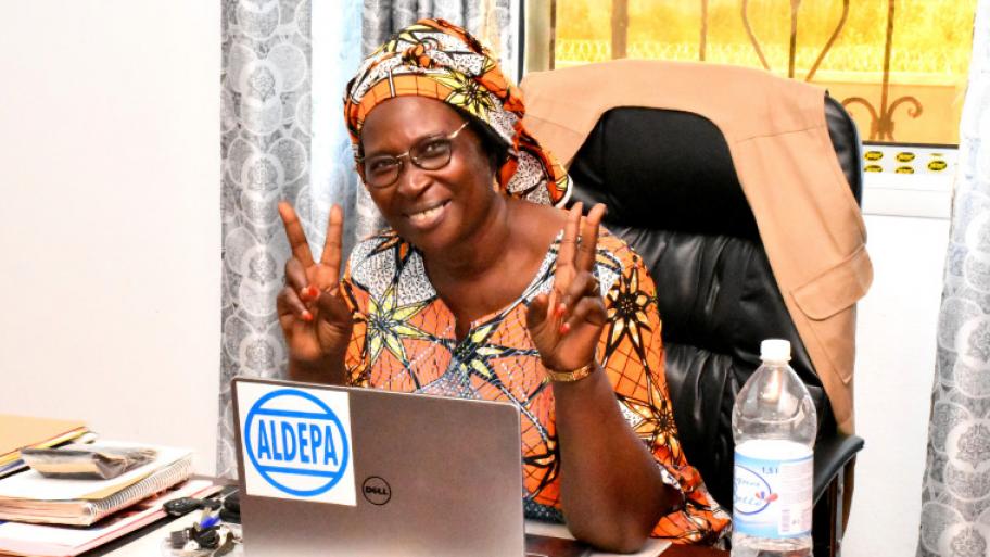 Die Gewinnerin des Alternativen Nobelpreises 2021 Marthe Wandou sitzt an einem Schreibtisch hinter ihrem Aufgeklappten Laptop, sie lächelt in die Kamera und bildet mit beiden Händen das Peace-Zeichen