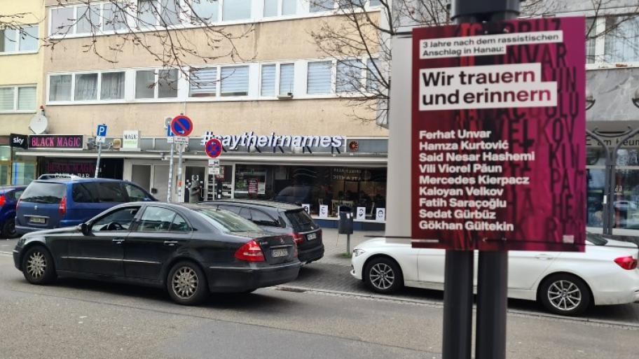 eine Straße in Hanau, von Autos befahren, im Hintergrund ein Laden/Cafè mit Namen #saytheirnames, im Vordergrund ein Plakat mit den Namen der Opfer des Anschlags von Hanau und dem Zusatz "3 Jahre nach dem Anschlag: Wir trauern und erinnern"