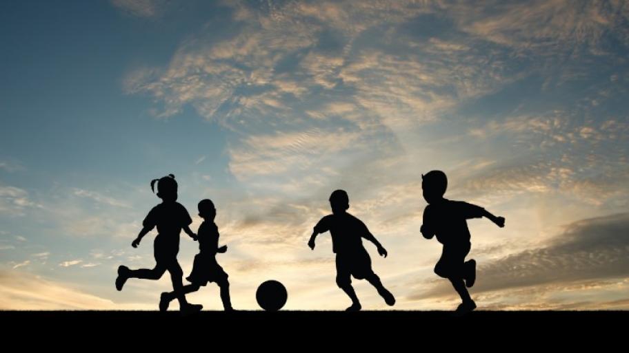 Drei Kinder spielen Fußball. Man sieht nur ihren Schatten und den blauen Himmel dahinter