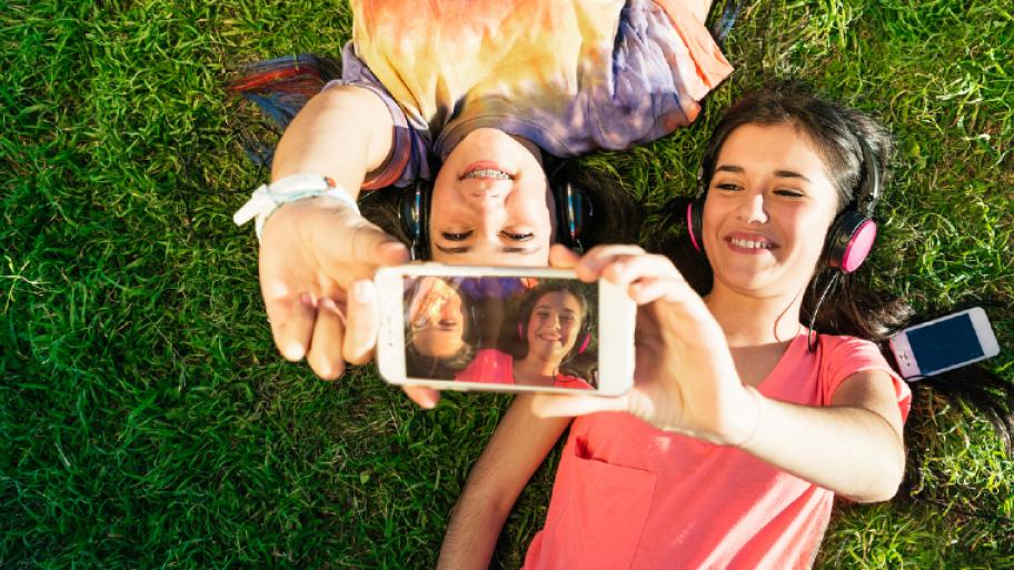 2 Mädchen mit Kopfhörern liegen auf der Wiese und machen ein Handy-Selfie