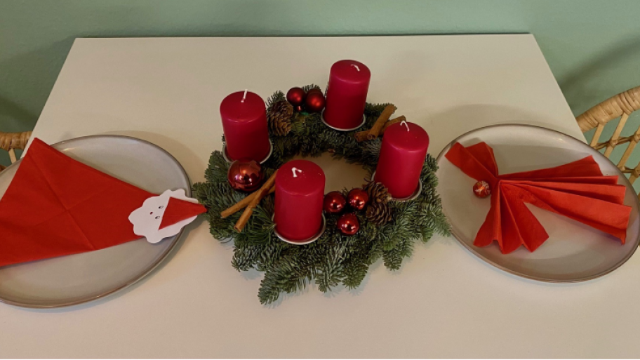 Zu sehen ist ein Tisch auf welchem ein Adventskranz liegt. Daneben liegen auf zwei Tellern zwei weihnachtlich gefaltete Servietten.
