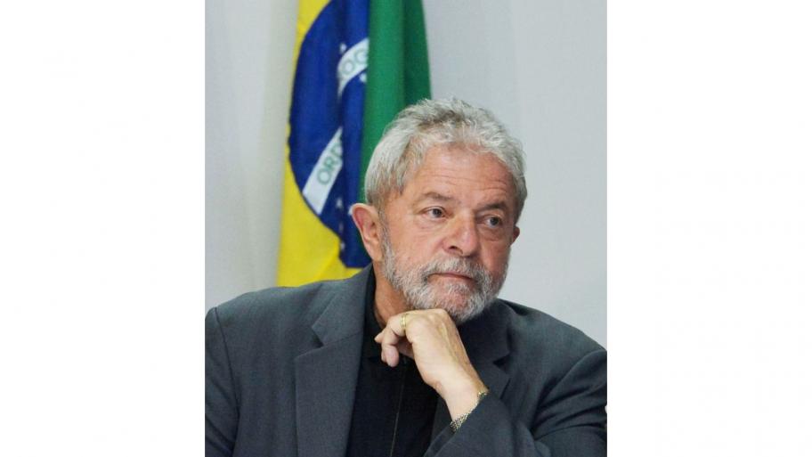 ein Porträt des brasilianischen Präsidenten Luiz Inácio Lula da Silva: er trägt einen grauen Anzug, stützt das Kinn auf die linke Hand, im Hintergrund die Flagge Brasiliens
