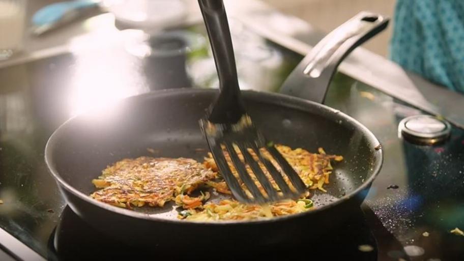 eine dunklen Pfanne steht auf einem dunklen Kochfeld, darin braten 3 Gemüsepuffer, einer von ihnen wird gerade mit einem schwarzen Pfannenwender gewendet