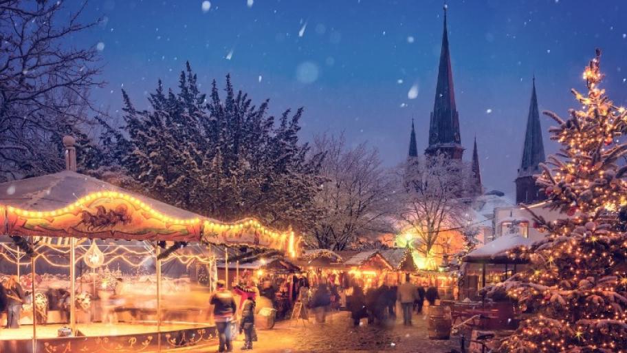 Beleuchteter Weihnachtsmarkt mit Karussell, Weihnachtsbaum und Holzhütten, im Hintergrund eine Kirche, es schneit