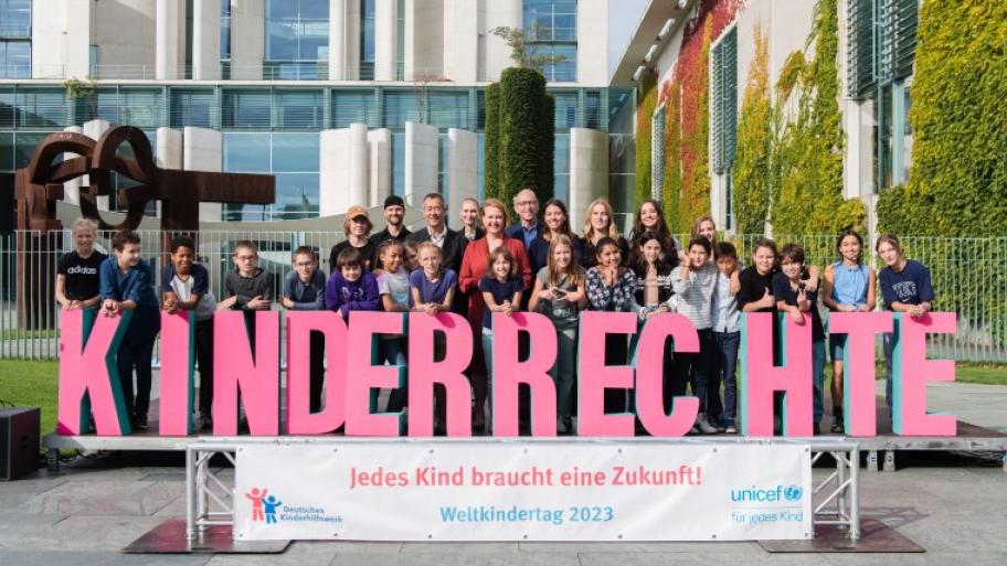 eine Gruppe Menschen steht auf einem Podest vor dem Bundeskanzleramt in Berlin, darunter Berliner Schüler*innen und Bundesfamilienministerin Lisa Paus, vor sich  das Wort "Kinderrechte" in großen rosa Buchstaben und auf einem Banner das Motto des Weltkindertages 2023 "Jedes Kind braucht eine Zukunft!"