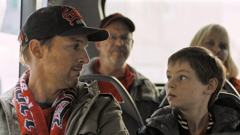 ein Mann und ein Kind sitzen im Bus. Der Mann sitzt links und guckt seinen jüngeren Sohn an, welcher rechts sitzt und ihn zurück angeguckt. Der Vater trägt eine Kappe und einen Fußball-Schal