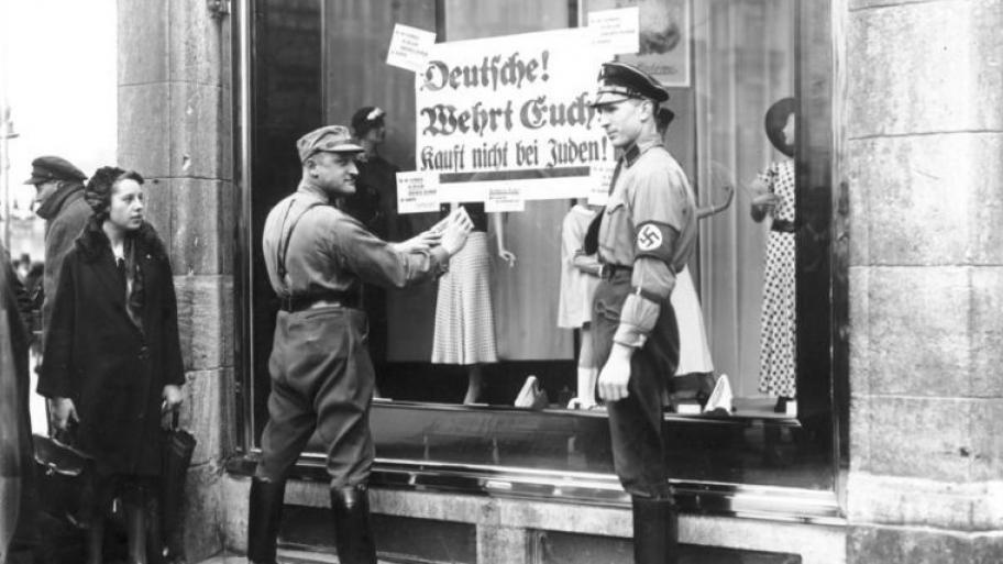 Schwarz-weiß Bild: zwei Nationalsozialisten bringen Schild an Schaufenster an. Darauf steht: "Deutsche wehrt euch. Kauft nicht bei Juden!"