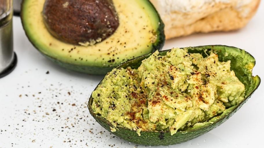 eine aufgeschnittene Avocado, eine Hälfte mit Kern, in der anderen Hälfte ist Avocado-Creme angerichtet, im Hintergrund ein Stück Weißbrot