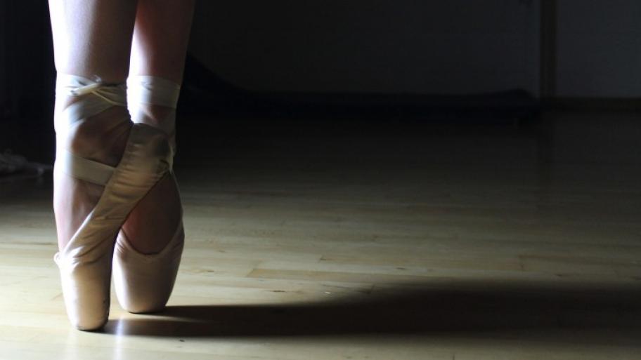 jemand tanzt Ballett in Spitzenschuhen, man sieht nur die Füße in den Schuhen, heller Boden, dunkler Hintergrund