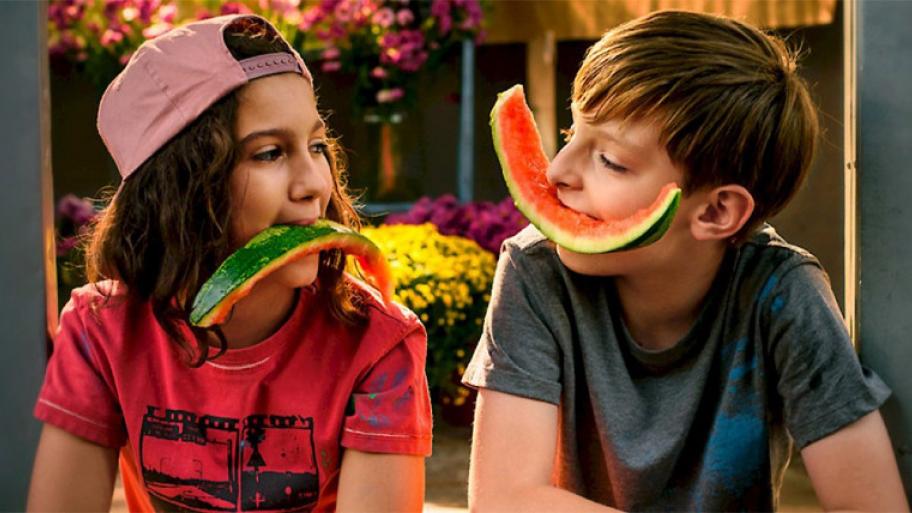 zwei Kinder sitzen sich gegenüber und schauen sich an, beide haben ein großes Stück Melonenschale quer im Mund
