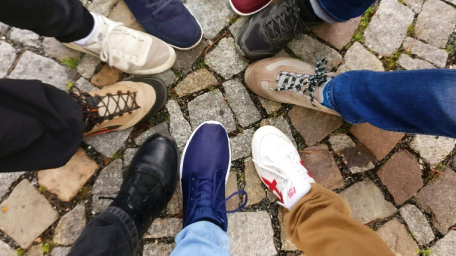 viele Füße in verschiedenen Schuhen finden sich kreisförmig zusammen, Untergrund gepflasterter Platz, von den Schuhträgern sieht man nur Ansätze des Hosenbeines