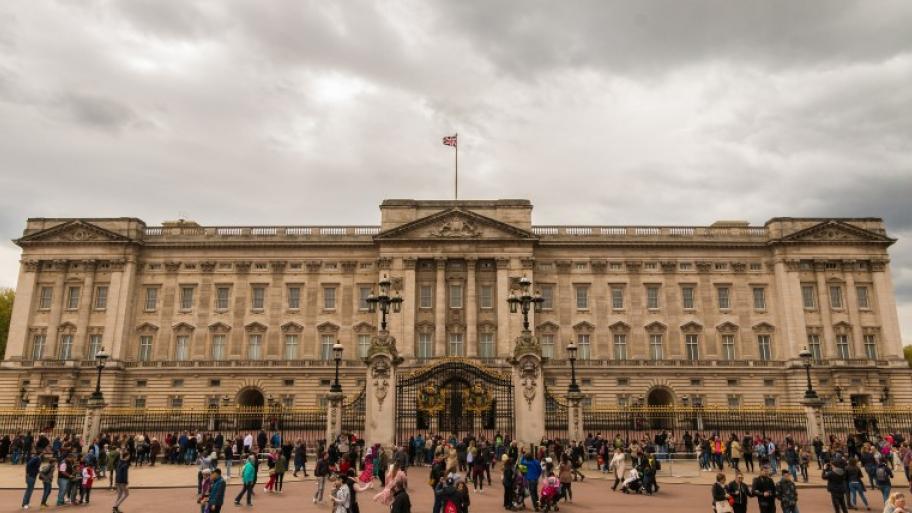 Frontansicht des Buckingham Palace in London, wolkiger Himmel im Hintergrund, davor einige Touristen