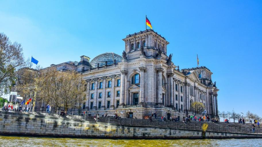 der Deutsche Bundestag (Reichstagsgebäude) von Wasserseite aus, ein paar Passanten davor, auf den Türmen wehen Deutschland- und Europa-Fahnen
