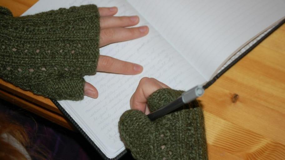 Kinderhände schreiben in grünen Wollstulpen mit Bleistift in der rechten Hand in ein geöffneten, liniertes Tagebuch