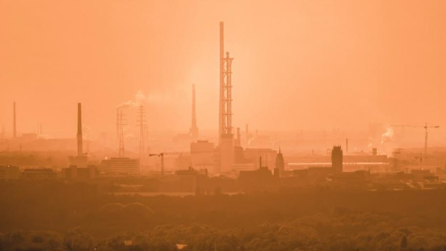 eine Stadtansicht mit vielen rauchenden Schornsteinen, im Hintergrund scheint die Sonne, die Luft ist orangefarben und sehr verqualmt