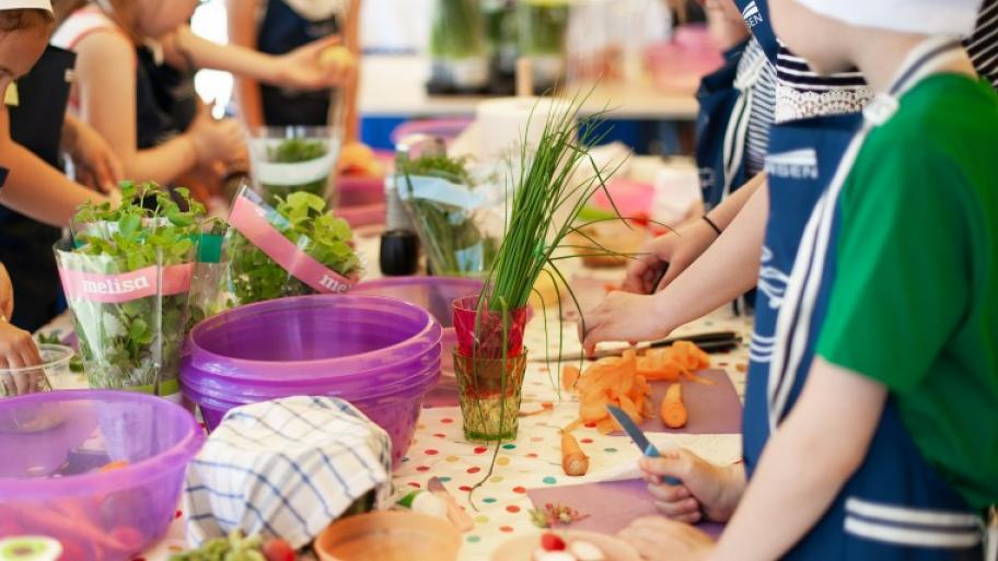 mehrere Kinder mit blauen Schürzen und weißen Kochmützen stehen um einen Tisch herum und schneiden frische Kräuter und Gemüse