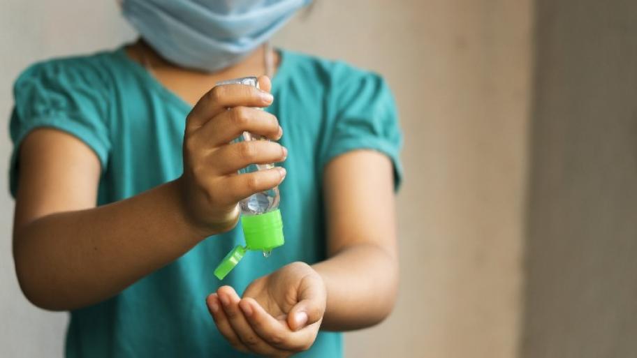 Kind mit medizinischer Maske vor dem Mund hält mit einer Hand Desinfektionsgel und lässt dieses in die andere Hand laufen