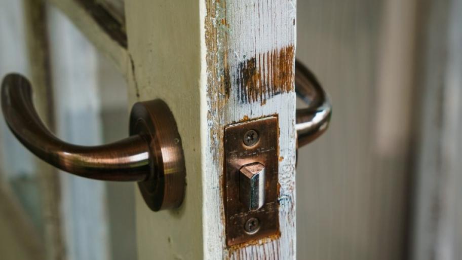 Großaufnahme des metallenen Türschlosses einer alten, weißen Holztür, daran auf beiden Seiten Metallklinken, die Tür ist geöffnet