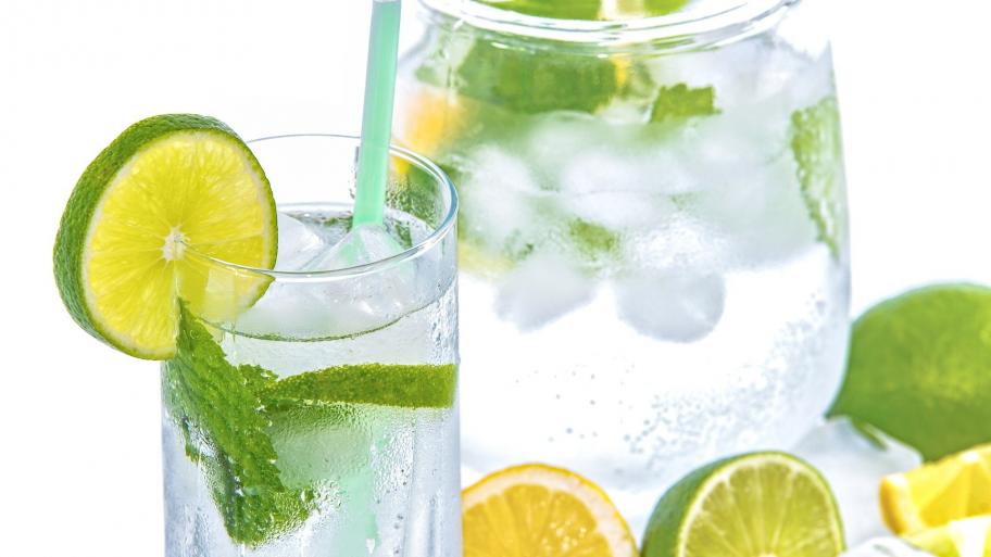 ein Glas und eine Karaffe gefüllt mit durchsichtiger Limonade, beide Gefäße mit Minze und Limette garniert, Limette auch drumherum als Dekoration, weißer Hintergrund
