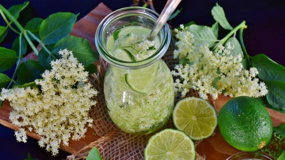 ein gedeckter Tisch, darauf eine Karaffe mit gelblicher Holunder-Limonade, daneben aufgeschnittene Limetten und weiße Holunderblüten