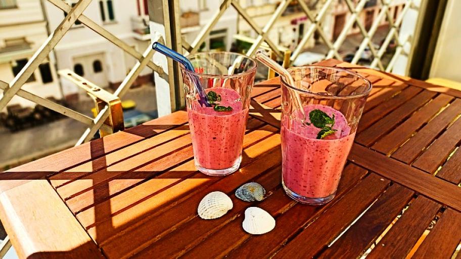 auf einem hölzernen Balkontisch zwei Gläser mit Strohhalm, gefüllt mit einem Rote-Beeren-Minze-Eisshake, als Deko 3 Muscheln