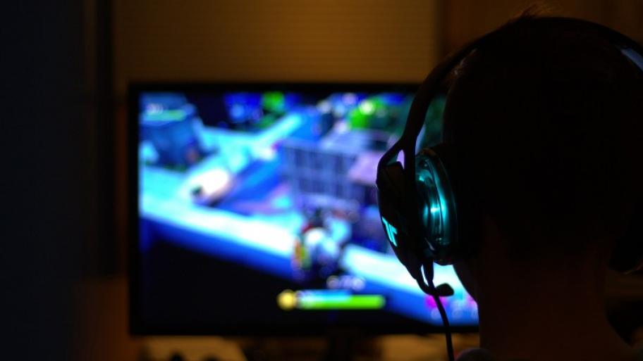 die schwarze Silhouette eines Kindes mit Kopfhörern von hinten, das vor einem leuchtenden Bildschirm sitzt, auf dem es das Videospiel "Fortnite" spielt