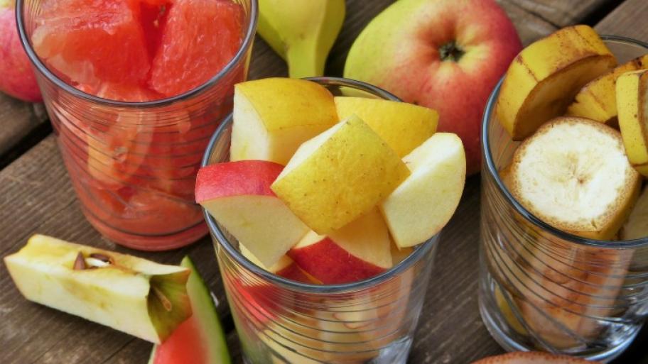 auf einem hölzernen Tisch stehen 3 Gläser gefüllt mit unterschiedlichen, geschnittenen Früchten, zum Beispiel Wassermelone, Apfel und Banane