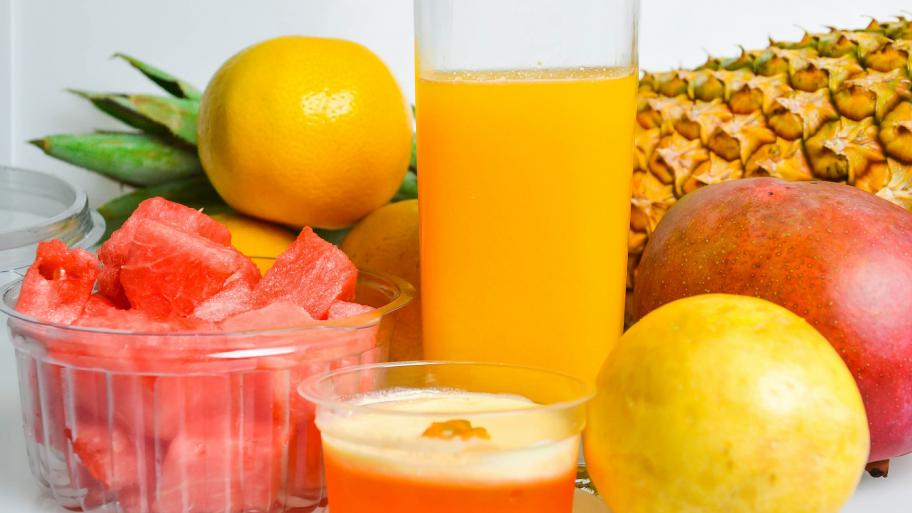 auf weißem Hintergrund eine Ananas, eine Mango, zwei Orangen, ein Glas mit orangenem Saft, eine durchsichtige Schale mit Wassermelonenstücken
