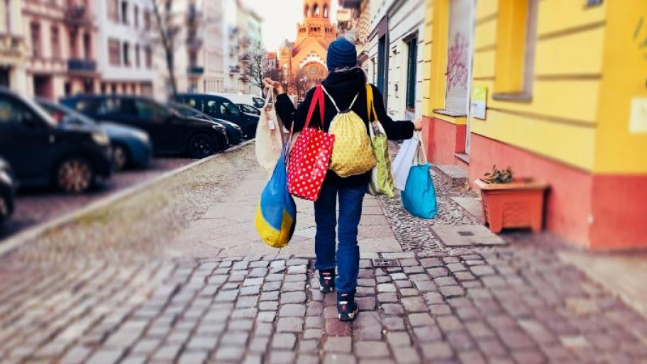 ein Kind geht überladen mit verschiedenen Einkaufstaschen und Beuteln den Bürgersteig entlang, im Hintergrund Autos und Häuserfassaden
