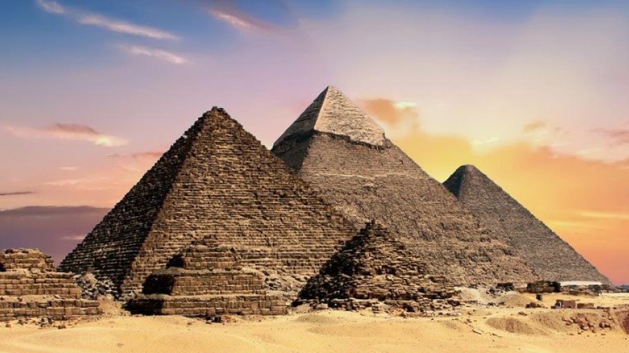 Zwei große sandfarbige Pyramdiden stehen im Mittelpunkt des Bildes. Darum verteilen sich noch einige Kleinere. Der Himmel leuchtet in lila, rosa, orangenen Farben während die strahlende Sonne hinter den Pyramiden verschwindet.