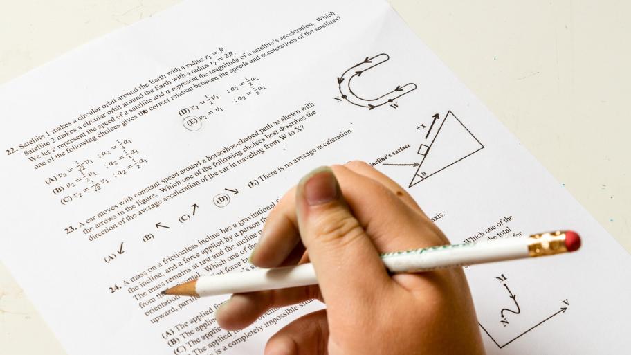 Eine Hand hält ein Stift. Dahinter liegt ein Arbeitsblatt mit Matheaufgaben. 
