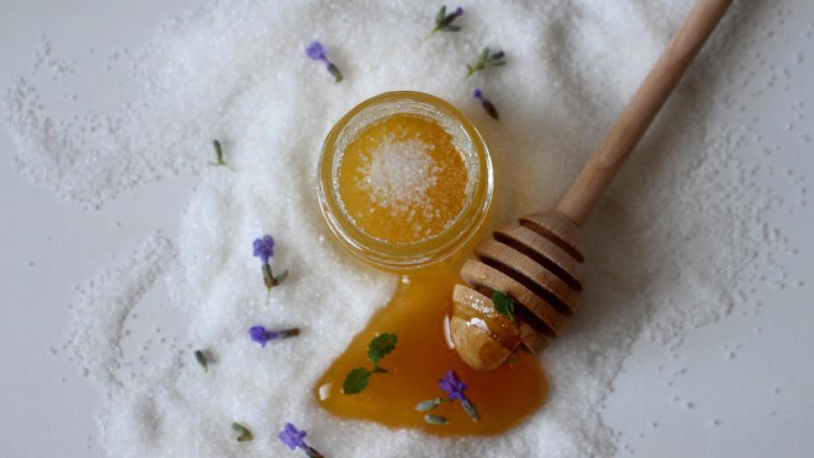 Kleines Glasgefäß mit Honig- und Zucker Scrub, Lavendelblüten liegen zerstreut um das Gefäß und ein Honiglöffel