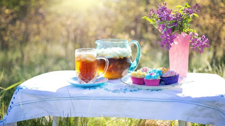 ein Tisch mit weißer Tischdecke steht auf einer Gräserwiese, darauf eine Vase mit Blumenstrauß, eine Karaffe und ein Glas gefüllt mit Eistee, ein großer Teller mit Cupcakes