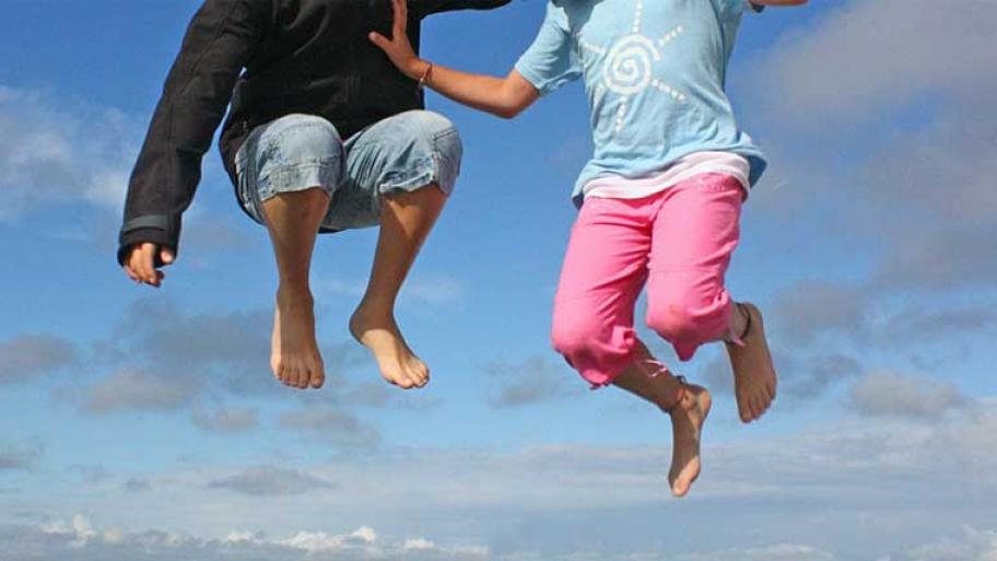 zwei Kinder springen hoch in die Luft, man sieht nur die Beine und die Oberkörper, nicht die Köpfe, im Hintergrund blauer Himmel und Wolken