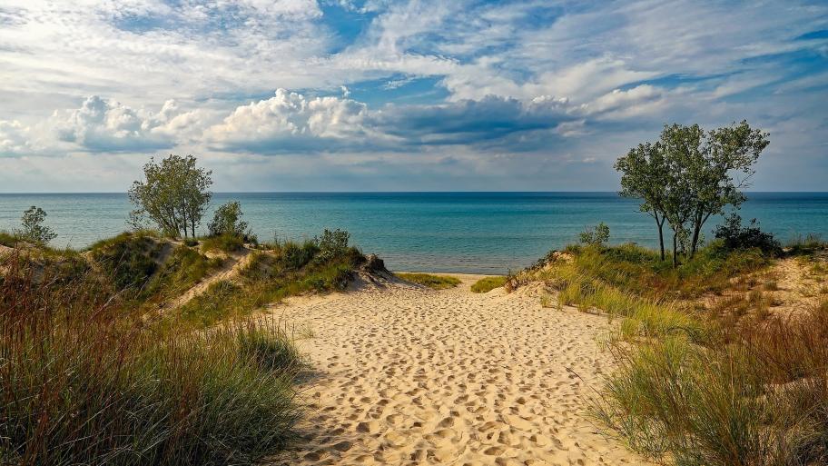 Sandweg zum Meer, dass schon im Hintergrund zu erkennen ist; rechts und links sind Dünen und andere Pflanzen; am Himmel sind Wolken