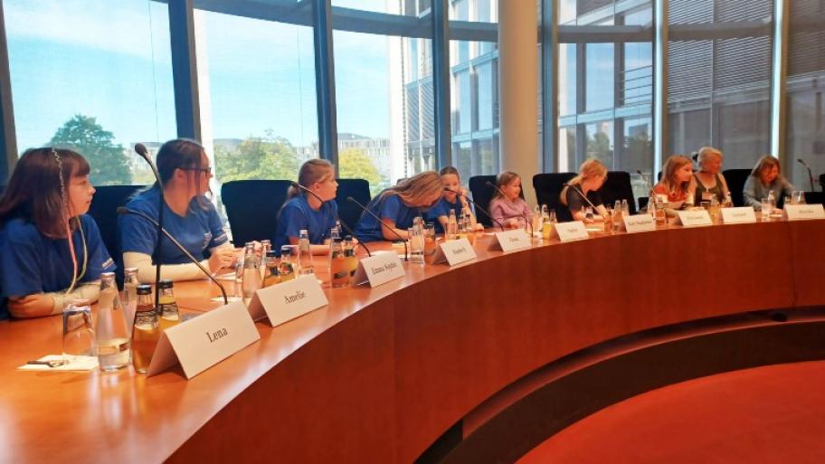 Kinder zu Besuch bei der Kinderkommission im Deutschen Bundestag: die Kinder sitzen an einem langen Tisch, vor sich Mikrofone und Namensschilder, im Hintergrund Fenster