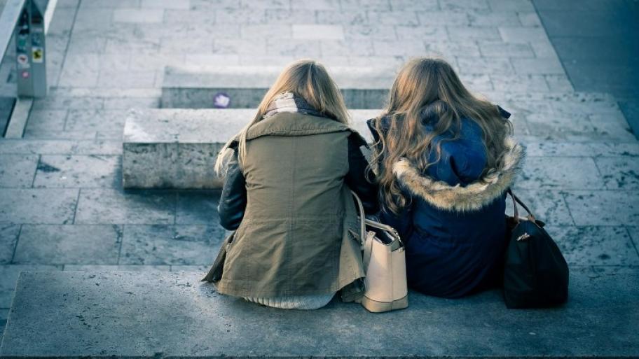 zwei Mädchen mit offenen Haaren von hinten, sie sitzen auf steinernen Stufen, rechts neben sich hat jede ihre Handtasche, sie tragen Jacken