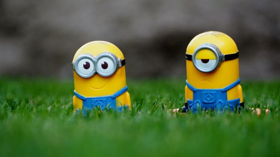 Zwei Minions sitzen im Gras, links Bob: lächelnd mit zwei Augen, rechts Stuart: grimmig, ein Auge 