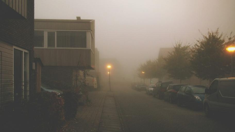 eine Straße im Nebel, links Häuser, rechts parkende Autos, zwei Straßenlaternen brennen
