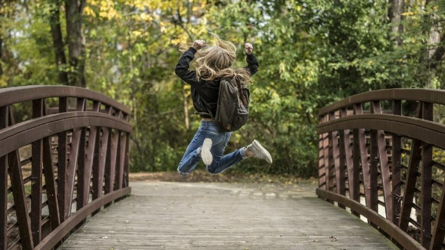 Ein Mädchen springt auf einer Holzbrücke freudig in die Luft. Im Hintergrund ist ein Wald zu sehen.