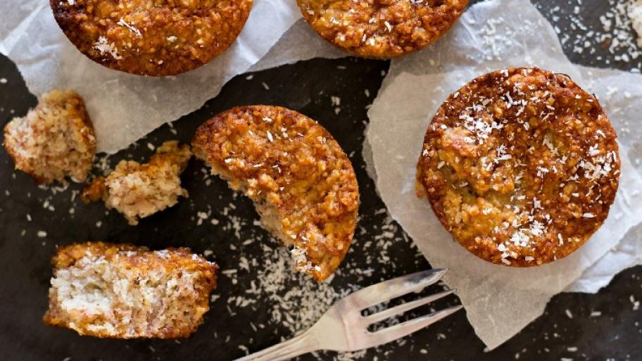auf einem dunklen Tisch liegen mehrere Bananenmuffins auf weißen Servietten mit Kokosraspeln bestreut, daneben eine silberne Gabel