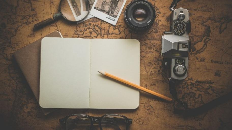 eine alte Landkarte, darauf liegen ein offenes Notizbuch mit Bleistift, eine Lupe, eine Brille, eine Kamera, zwei Fotos
