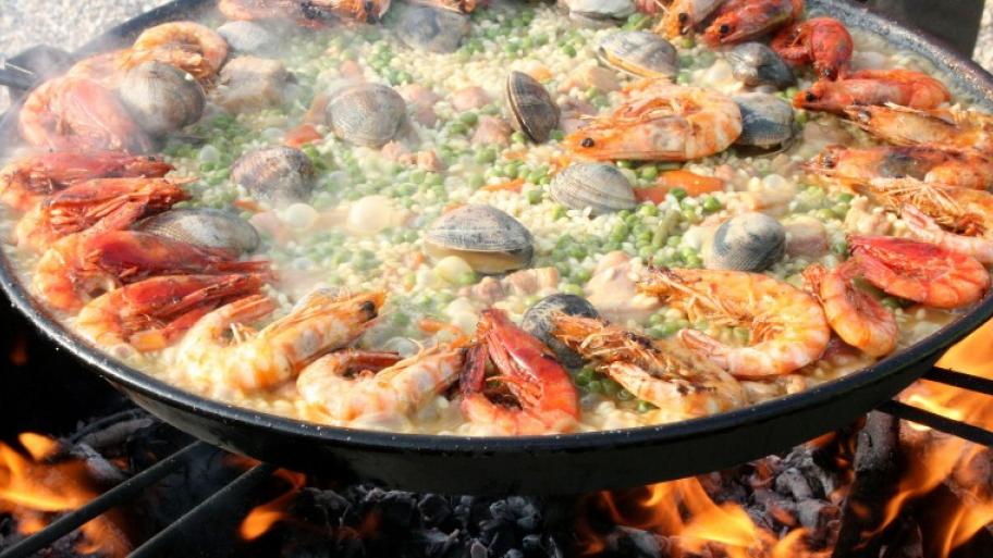 auf einer Feuerstelle steht eine sehr große, gusseiserne Paella-Pfanne, gefüllt mit Paella aus Reis und Erbsen, garniert mit Muscheln und garnelen