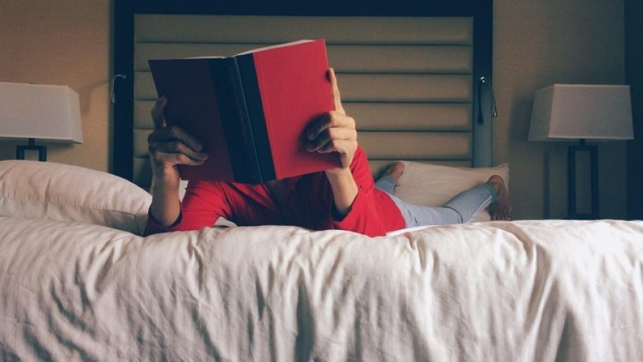 eine Person in rotem Pulli und grauer Hose liegt bäuchlings auf einem Bett, hält in den Händen ein rotes, aufgeschlagenes Buch, das Gesicht verschwindet dahinter