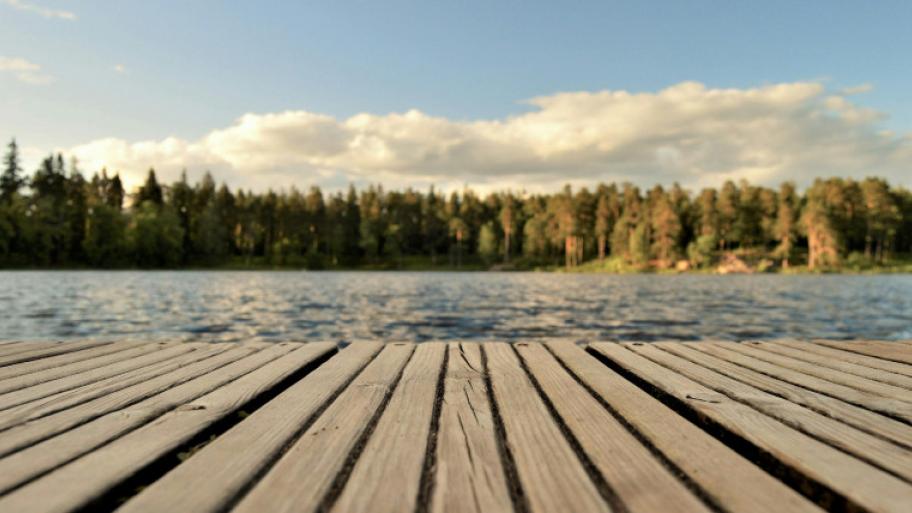Ein Steg über einem See. Am Rand des Sees steht ein Wald. Darüber blauer Himmel mit Wolken.