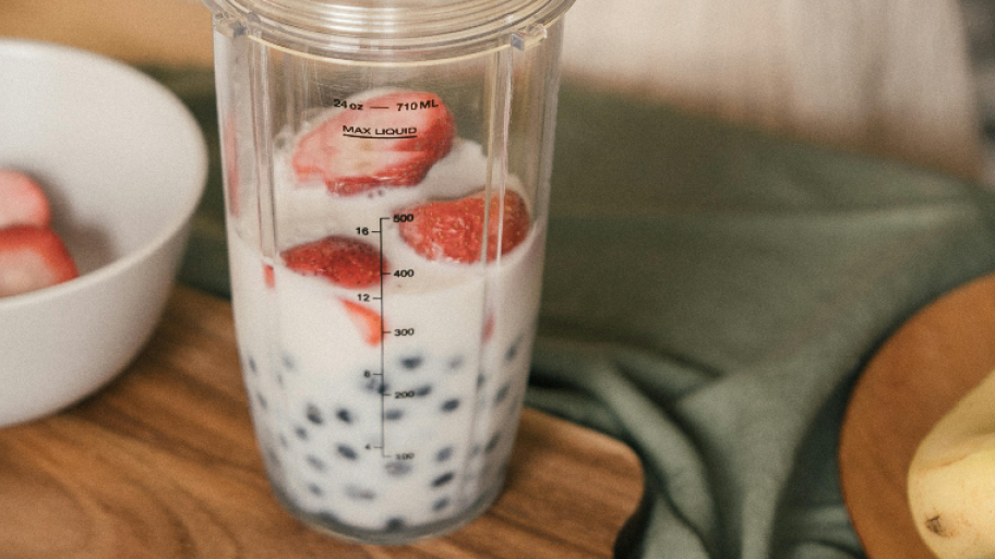 Smoothie Mixer gefüllt mit Milch, Blaubeeren und Erdbeeren, daneben steht eine weiße Schale mit aufgeschnittenen Erdbeeren auf einem Holzbrett