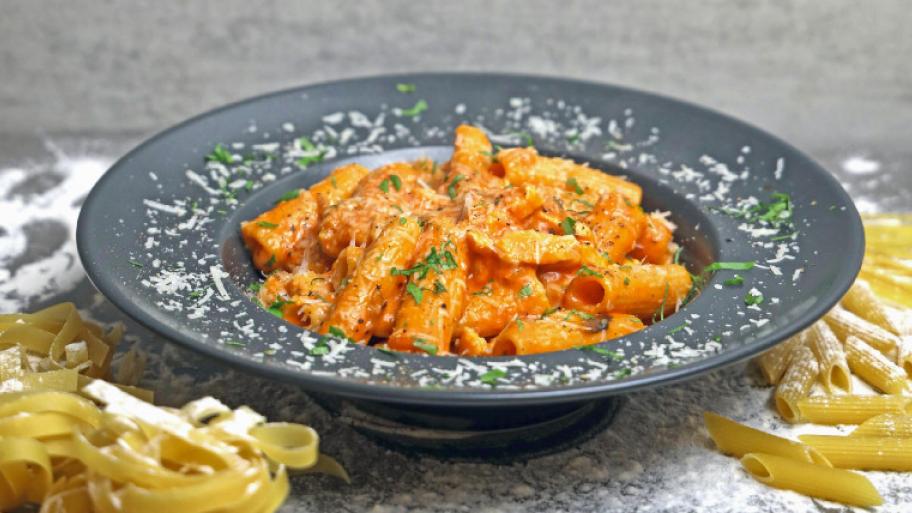 Dunkelgrauer Teller mit Nudeln in roter Soße. Darüber Parmesan und grüne Kräuter.
