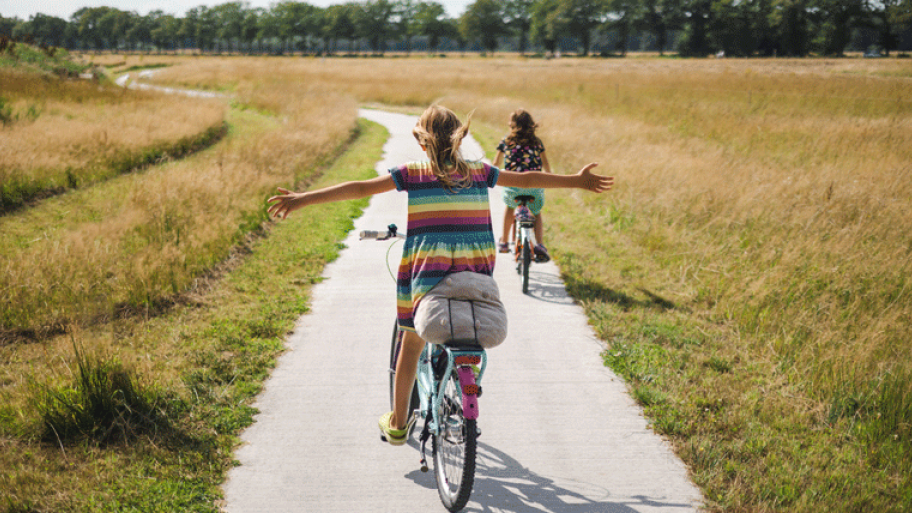 zwei Kinder auf dem Fahrrad, eins davon freihändig, es ist Sommer, links und rechts ist Wiese