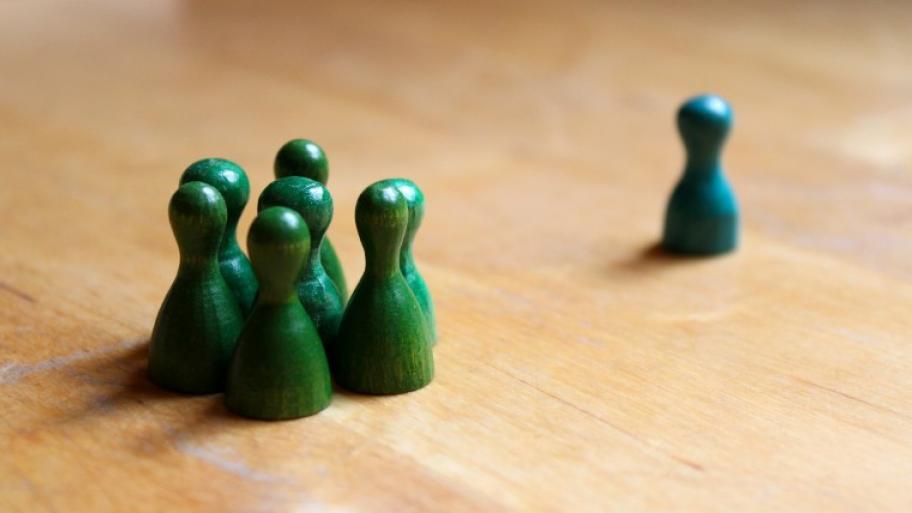 auf einem Holztisch steht eine Gruppe von 7 grünen, kleinen Spielfiguren einer einzelnen, blauen Spielfigur gegenüber
