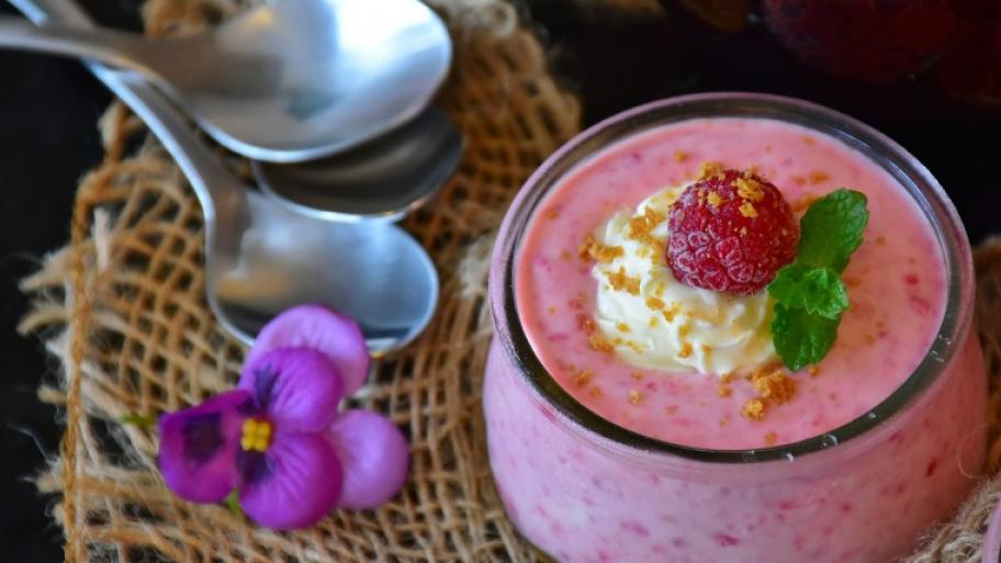 auf einem geflochtenen Bast-Untersetzer ein Glas gefüllt mit Himbeer-Quark, daneben zwei Esslöffel und eine pinke Blüte